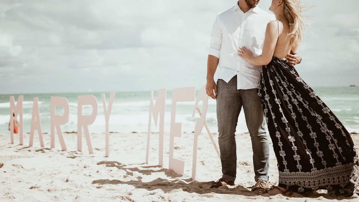 couple standing on marry me freestanding on beach